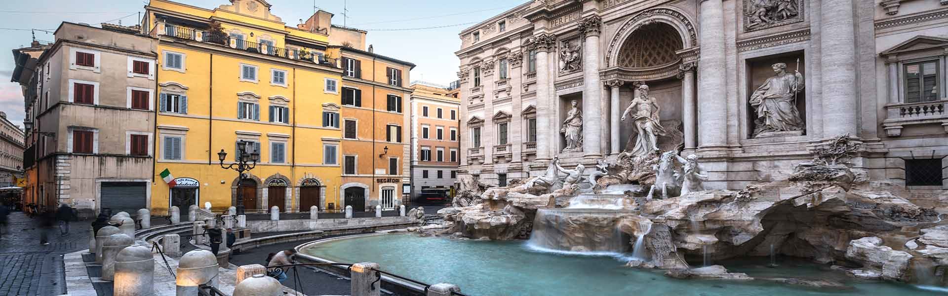 Seitenansicht des Trevi-Brunnens. Auf der rechten Seite sieht man den Brunnen mit seinem kristallklaren blauen Wasser und direkt dahinter den Palazzo Poli und sein Mauerwerk. Auf der linken Seite befinden sich mehrere orangefarbene Gebäude.