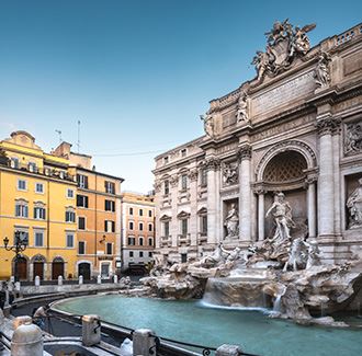 Seitenansicht des Trevi-Brunnens. Auf der rechten Seite sieht man den Brunnen mit seinem kristallklaren blauen Wasser und direkt dahinter den Palazzo Poli und sein Mauerwerk. Auf der linken Seite befinden sich mehrere orangefarbene Gebäude.